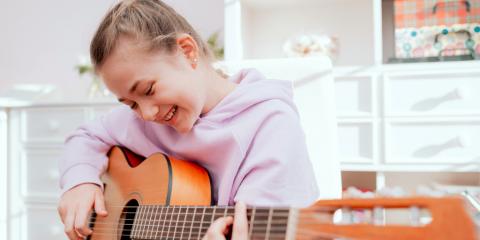 Smiling Caucasian teenage girl enjoys her free time playing acoustic guitar while sitting on a chair in her bright room.